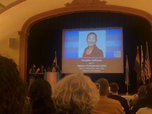 In a crowded auditorium, a screen at the front shows a smiling Korean name with her name and details below. At a podium, a tall, slender, elderly Black woman is reading about Maddie.