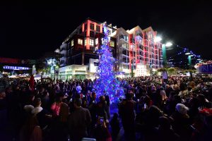 A crowd of people surrounding a light blue christmas tree with apartment buildings in the back