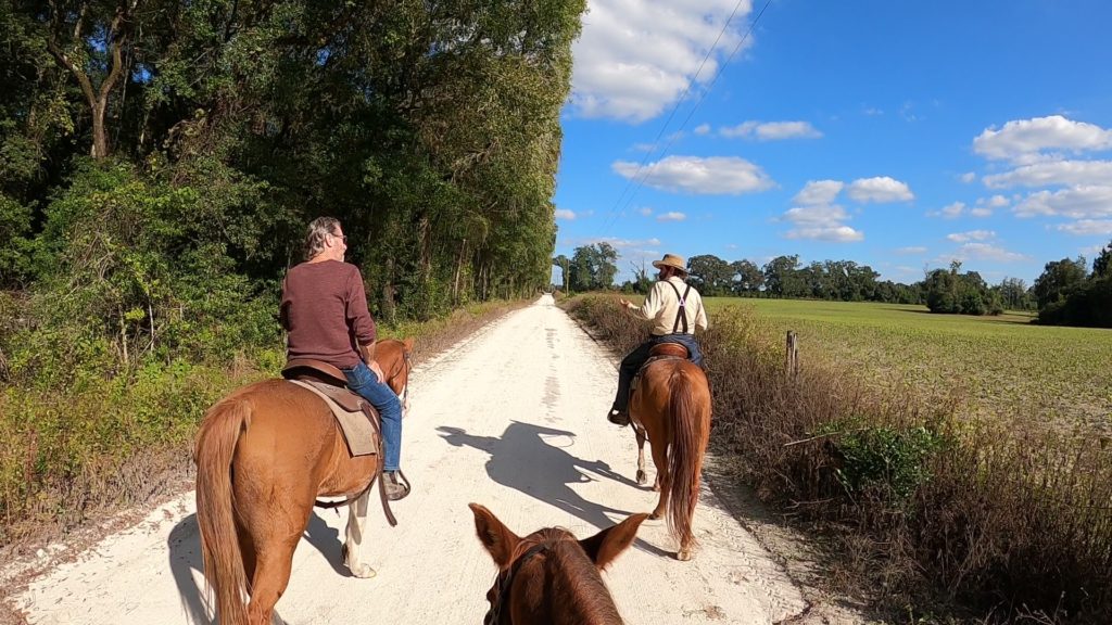 Two men on horses and the ears of a third horse...on a wide dirt road beside a green field with blue sky filled with cotton-candy clouds