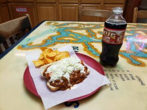 Loaded hot dog, with chili and slaw, BBQ chips and a root beer on a table