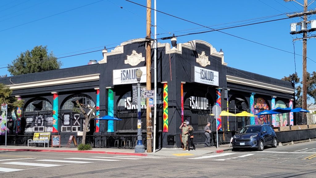 Black building with colorful murals on street corner, sign reads "Saluid!" Salud! Tacos, Barrio Logan Restaurants.