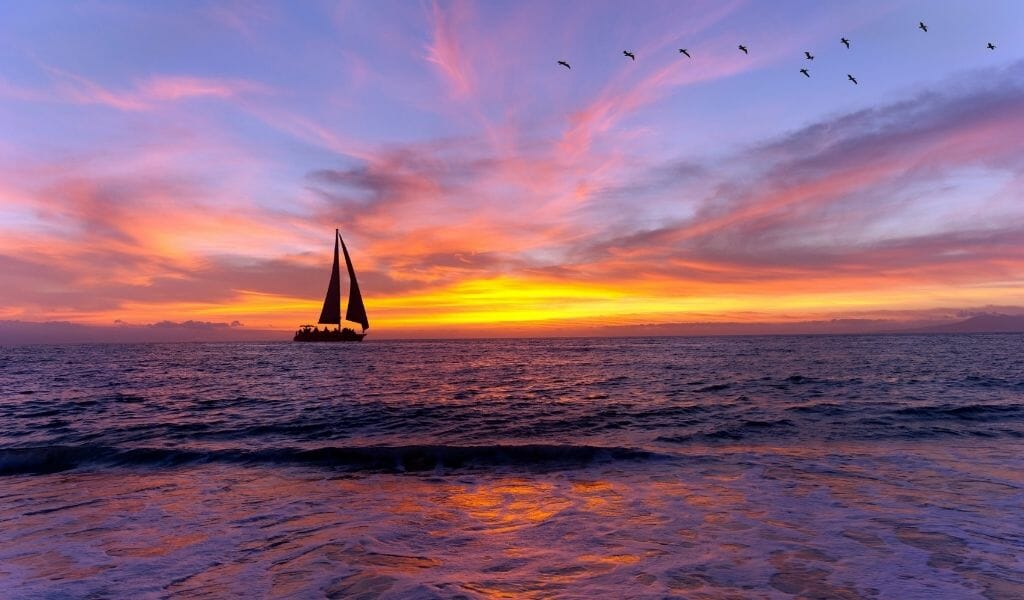 Sail boat near coast during sunset with pink clouds, purple sky, and orange sun