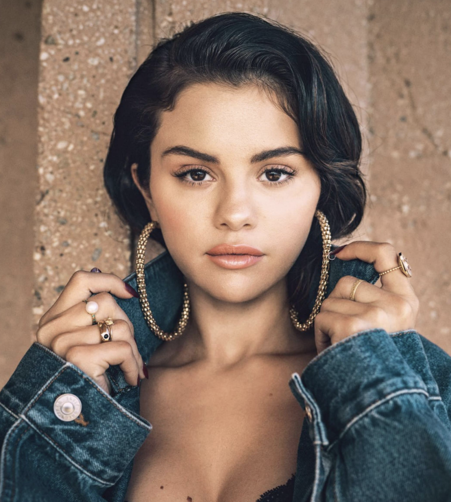 Headshot of Selena Gomez with large gold hoops, gold jewelry, and a jean jacket