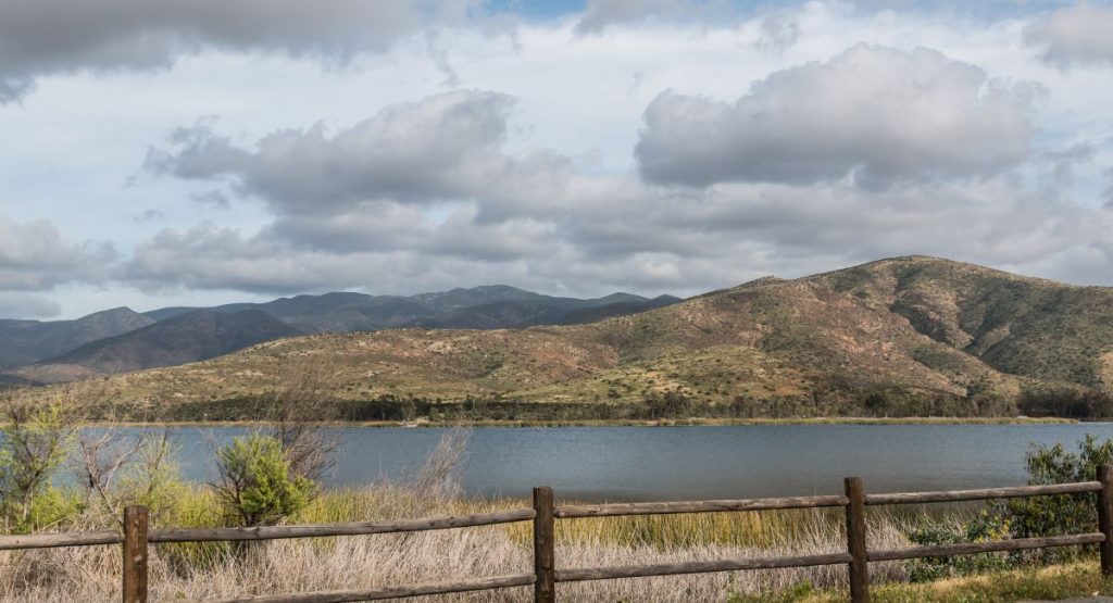 Wooden fence along path in front of large lake with small mountains in background on cloudy day. Otay Lake, San Diego lakes.