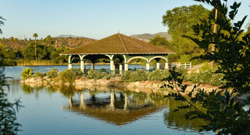 Large covered gazebo on stone outcropping covered in greenery extending into calm lake, with distant mountain and palm tree views. Lindo Lake, lakes in San Diego.