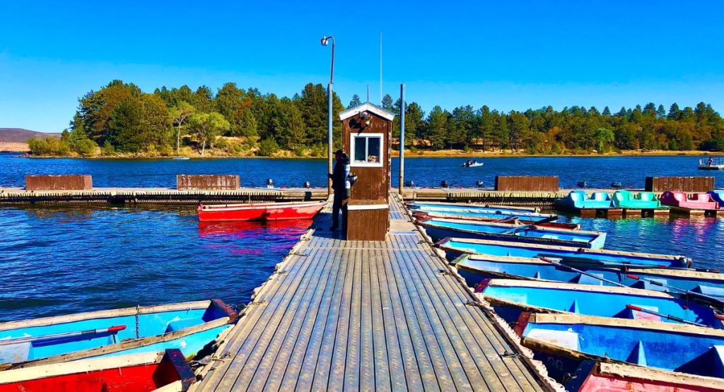 Narrow wooden dock lined with small wooden rental boats on blue lake with trees and sunny skies in background. Lake Cuyamaca, California.