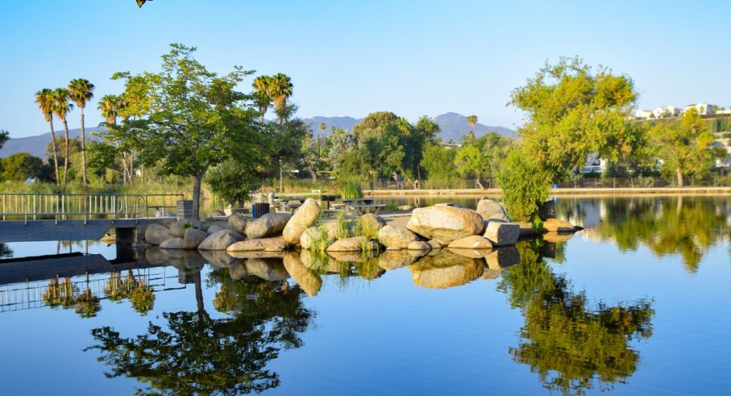 Large boulders, trees, and small bridge surrounded picnic benches on edge of calm lake with distant mountains in background on blue sunny day. Santee Lakes, San Diego lakes.