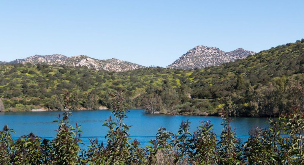 Blue lake sitting below green rolling hills covered in low shrubs with desert-like rocky mountains in background on sunny day. Lake Jennings, lakes in San Diego.