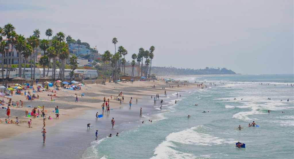 Sandy beach covered in beach-goers with bright umbrellas and water toys, palm trees and buildings line the beach opposite of the Pacific Ocean. Carlsbad Beach.