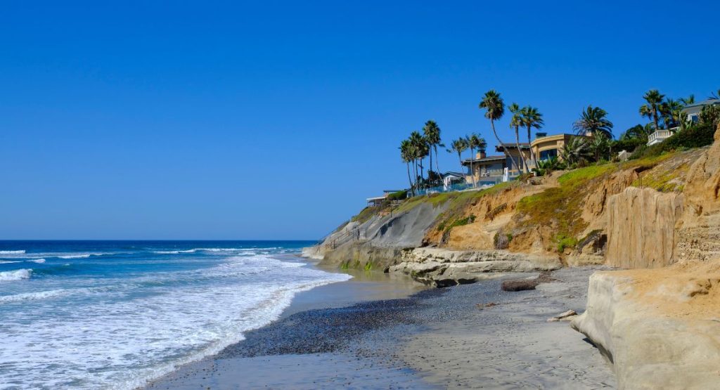Waves breaking on sandy beach next to hill with large homes and palm trees on sunny summer day. Terramar Beach, Carlsbad Beaches.