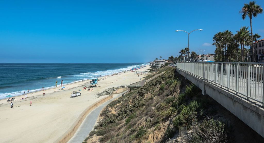 Wide sidewalk lined with buildings and palm trees overlooking wide white sand beach dotted with beachgoers on Pacific Ocean on cloudless day. Carlsbad State Beach. Carlsbad attractions.