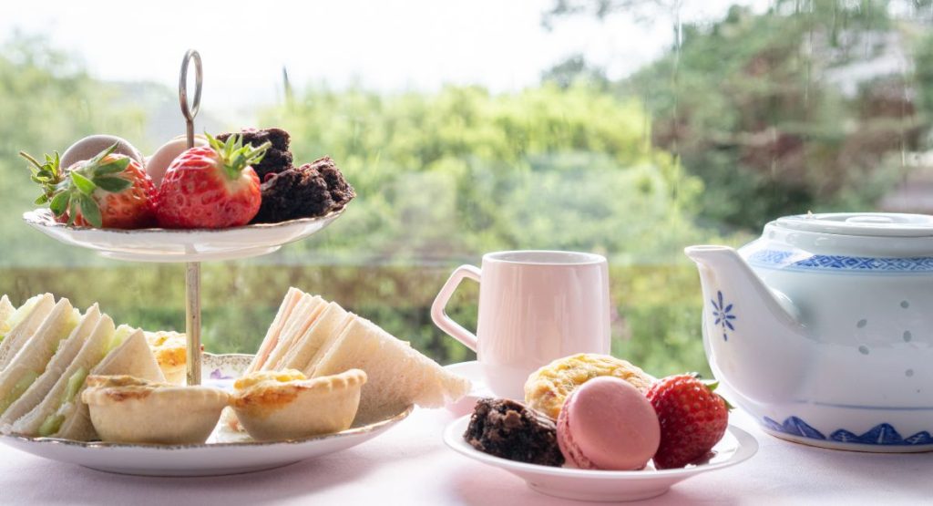 Close up shot of tiered tray of finger food and fruit, tea cup, and blue floral China tea pot in natural light in front of window with green trees in background. Afternoon Tea San Diego.