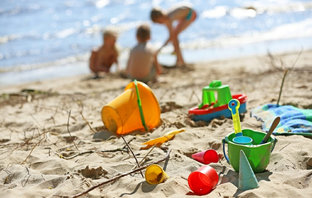 colorful beach toys and buckets laying in the sand with three kids playing on the beach in the background