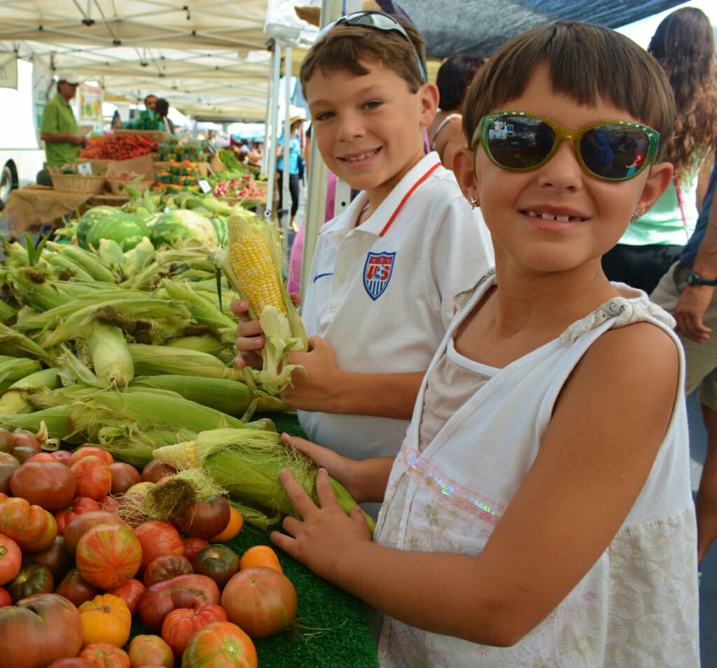 A young girl with white dress and sunglasses in front of a young boy in white tshirt - both standing next to a vegetable booth at the farmers market in san diego