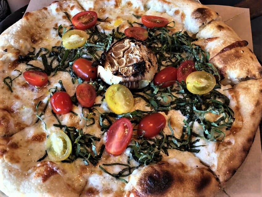 Closeup of pizza with goat cheese, halved cherry tomatoes and stripes of basil at Goat & Vine Temecula Restaurant