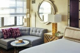 Hotel room with bed, corner sectional sofa in grey and a round gold mirror