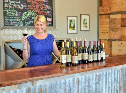 Winetasting Manager Mel standing behind the counter holding a glass of wine and lots of wine bottles on the counter