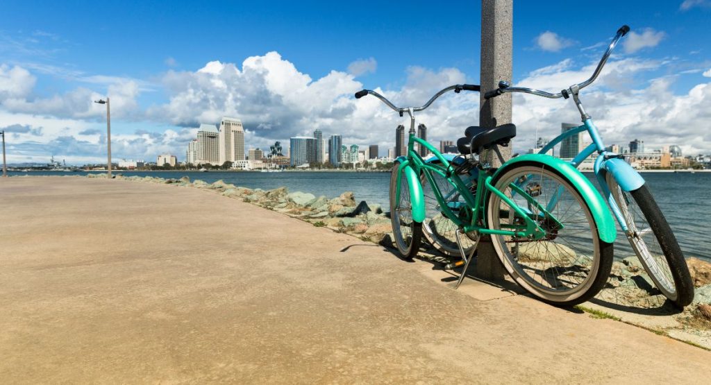 Two blue cruiser bikes propped up on concrete promenade overlooking San Diego Bay and San Diego skyline. Bike the Bay San Diego, August events in San Diego.