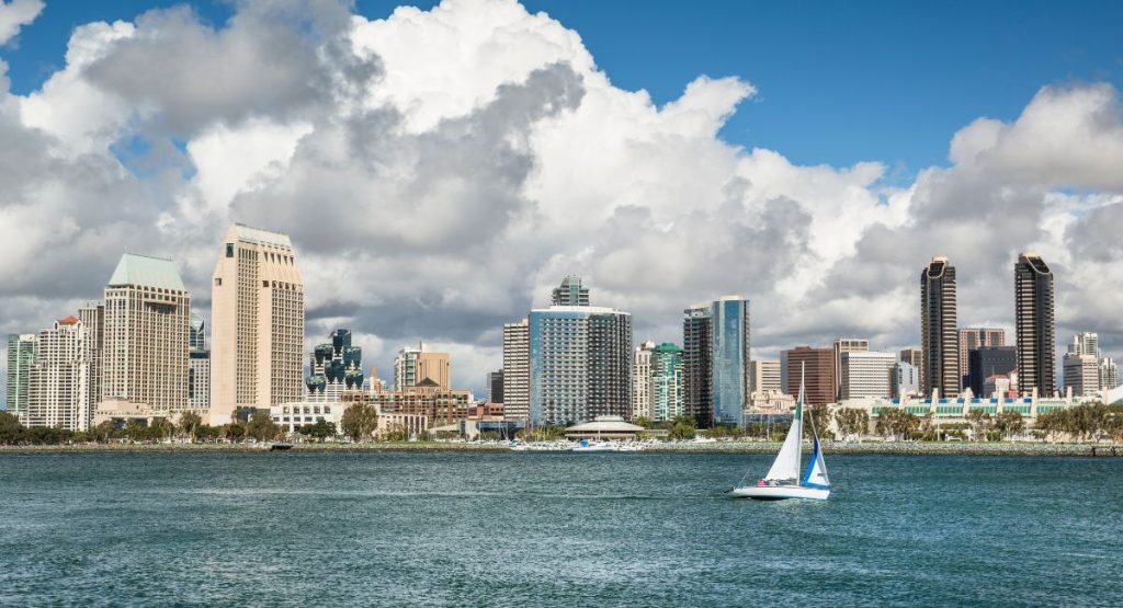 Small sailboat sailing calm waters with San Diego skyline in background on sunny day. Sailboat tours San Diego.