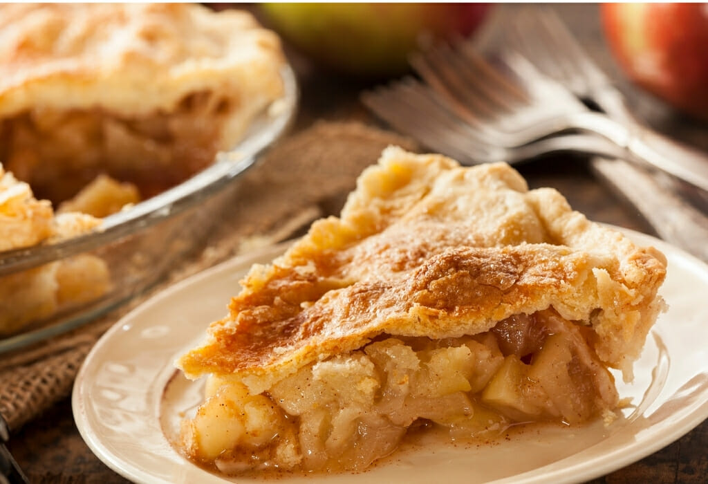 Large piece of apple pie on white plate in front of pie tin sitting on a wooden table with forks and apples in the background - Julian Apple Pie