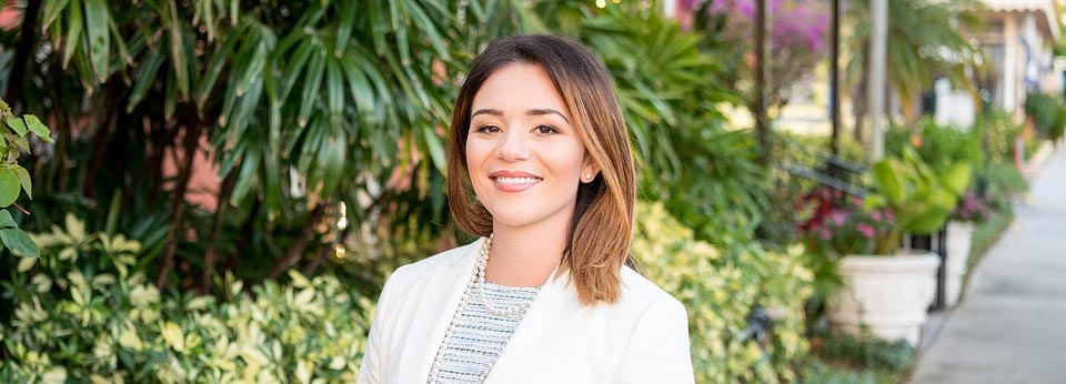 Lady with jacket. Foliage behind her. Sidewalk. Tree trunks. Planters.