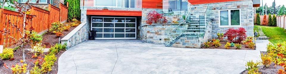 Front of house. Driveway. Landscaping, bushes. Wooden fencing. Stone exterior. Garage door. Steps. Windows. Small trees.