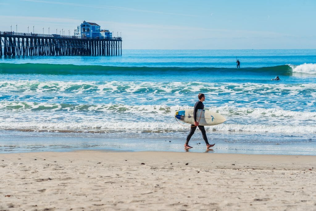 surfer with surfboard walking on the beach with Oceanside Pier in the background