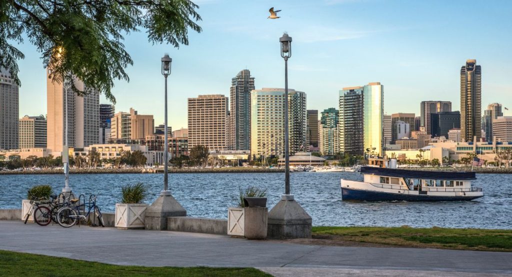 Sidewalk and grassy lawn of promenade overlooking San Diego Bay, ferry, and San Diego skyline during early evening on warm day. Coronado Ferry Landing Walkway.