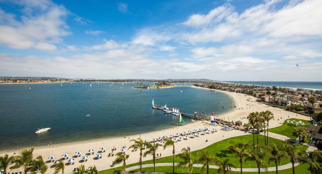 Hook shaped white sand beach with boardwalk pier, lounge chairs and umbrellas, and palm trees on sunny summer day. Mission Beach, snorkeling in san diego.