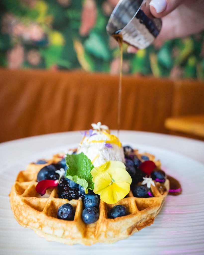 A waffle topped with an abundance of berries and edible flowers has syrup being poured over it delicately