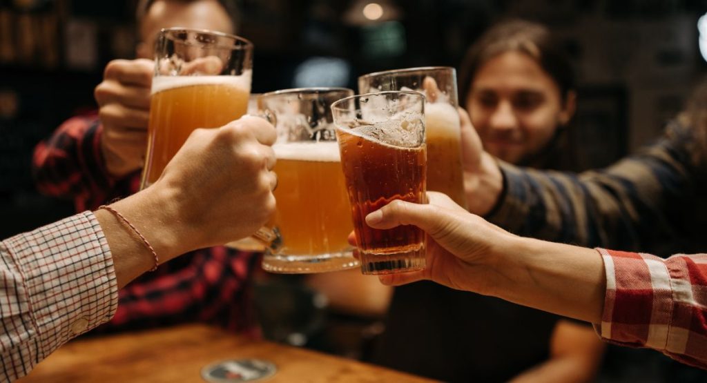 Group of friends smiling and cheersing large glasses of craft beer in dimly lit bar. Brewery tours of San Diego