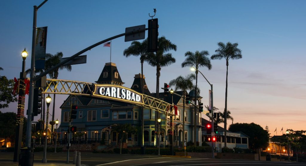 Large, backlit sign spanning street, reading "Carlsbad" in background are silhouettes of buildings and palm trees during late sunset. Carlsbad Village. What to do in Carlsbad.