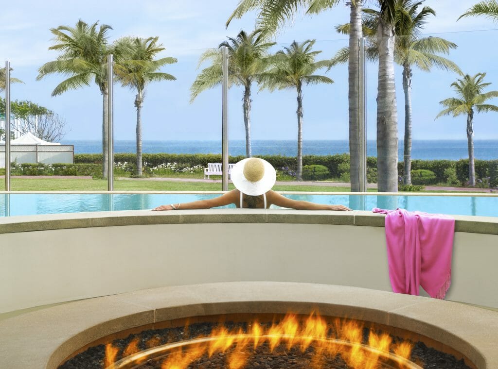 Gas firepit in the foreground. Woman sitting in pool with white hat, palm trees and Pacific Ocean in the background - Coronado Island Hotels - Hotel Del Coronado