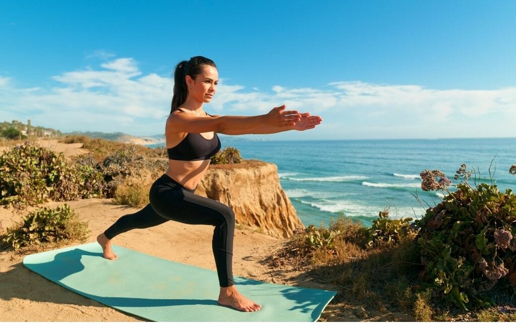 Things To Do in Dana Point-woman doing yoga warrior pose on a turquoise yoga mat on a cliff overlooking the ocean