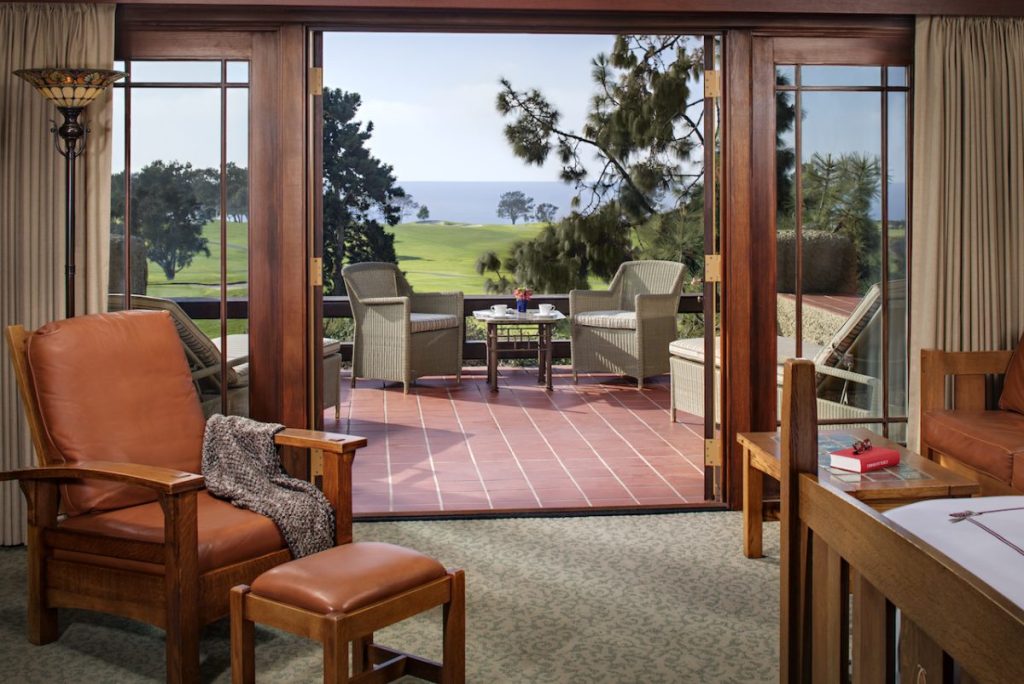 Luxury hotel room with leather sitting area and large outdoor balcony with wicker furniture, balcony overlooks golf course and distant ocean on sunny day. The Lodge at Torrey Pines.