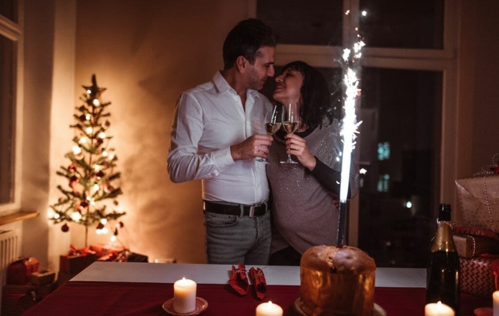 Couple toasting with champagne with cake decorated with firework sparkles and christmas tree in the background 