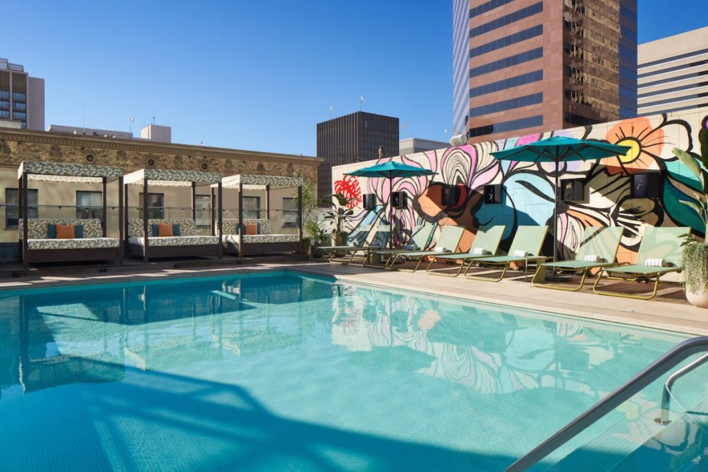 Rooftop pool area with large pool surrounded by cabanas and lounge chair under umbrellas, as well as colorful mural wall, tall city buildings and blue sky in background. Kimpton Alma, romantic Hotels in San Diego.