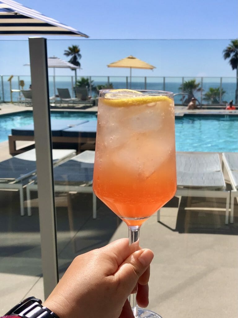 Orange drink held in front of pool at Blue and white macrame ceiling art at The Seabird Resort Oceanside