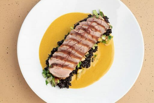 In a pool of silky yellow sauce, a perfect duck breast is slices and places over greens on a white round plate