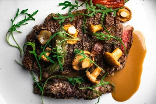 A steak on a white plate with greens and mushrooms scattered over it