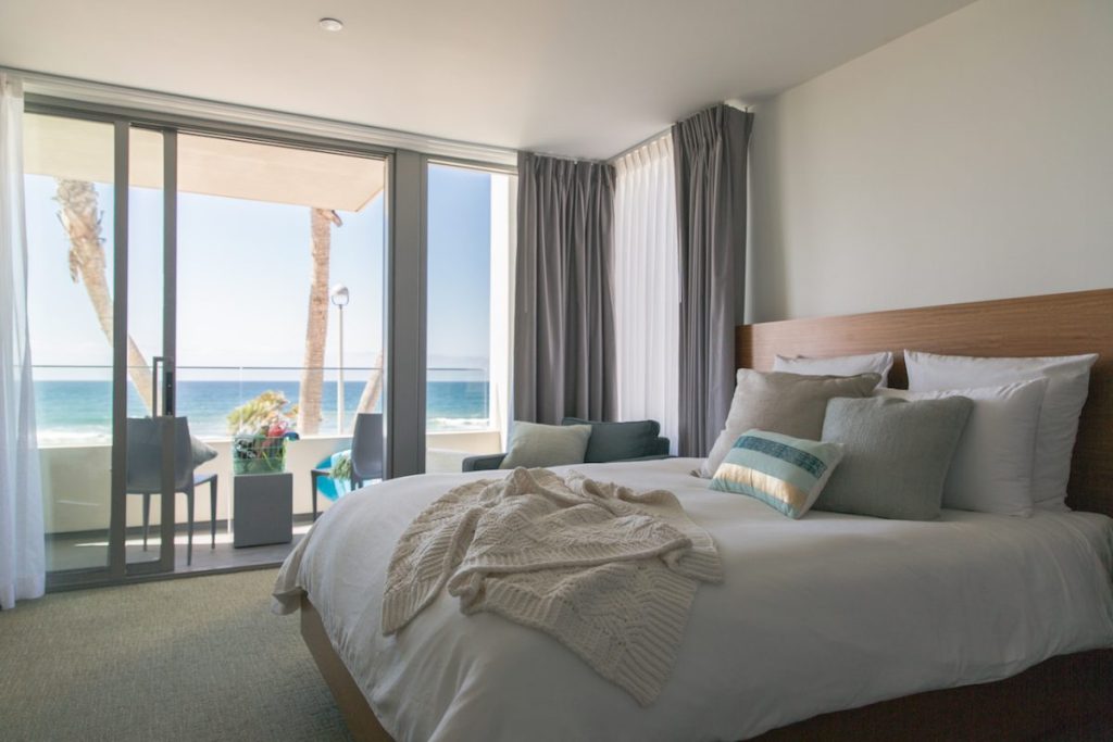 Photo of hotel room with king size bed with white linens, colorful pillows, and large balcony overlooking ocean views on sunny summer day. Tower 23, Best hotels in San Diego for Couple.