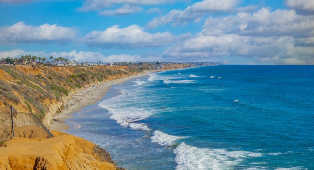 Blue water of the Pacific Ocean breaking on white sand beach next to sandy beach cliffs lined with palm trees on sunny day. North Ponto Beach, Carlsbad Beaches.