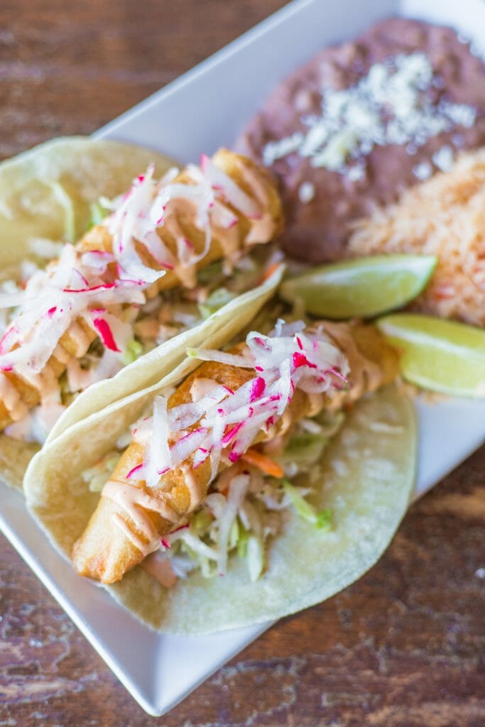 2 Fish Tacos with a side of rice and refried beans on a rectangular white plate