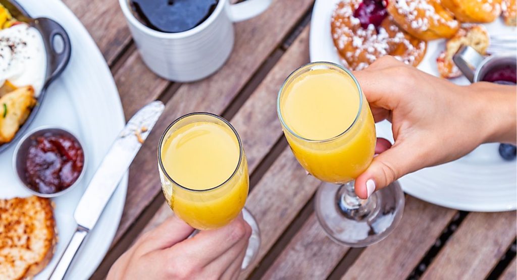 Two women holding mimosas in champagne glasses with plates of brunch food and coffee on table in background. Bottomless Mimosas in San Diego.