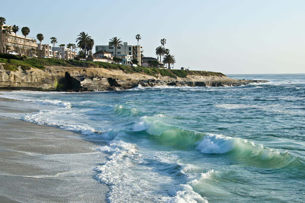 Water crashing onto the sand of the La Jolla shores with cliffs filled with buildings and greenery in the background