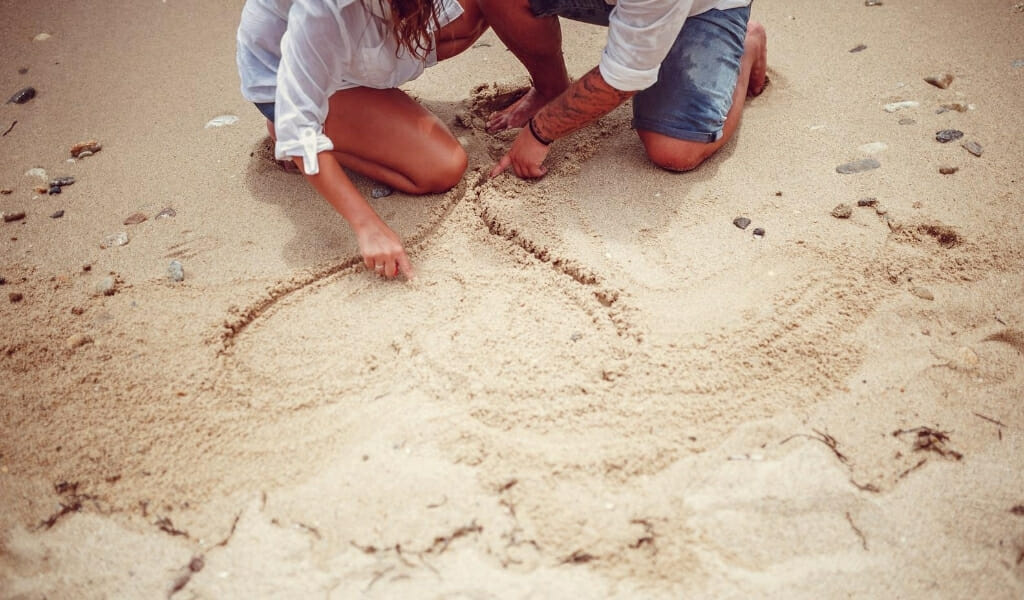 Partially visible man and woman kneeling the sand on the top edge of the photo, drawing a heart in the sand with their finger - Date Ideas San Diego - SanDiegoExplorer