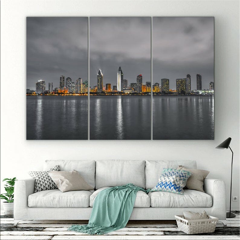 Photo of San Diego skyline split in three with grey, moody weather, accent color orange. It hangs in a living room with white couch on the wall below the pictures.