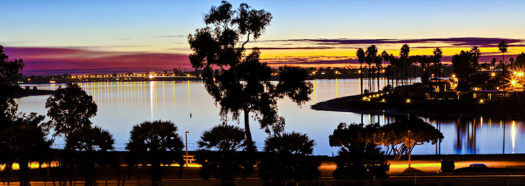 The sun sets of the water, creating hues of purple, blue, and yellow along the sky while the dark outlines of the land and trees stand stark against the colorful canvas