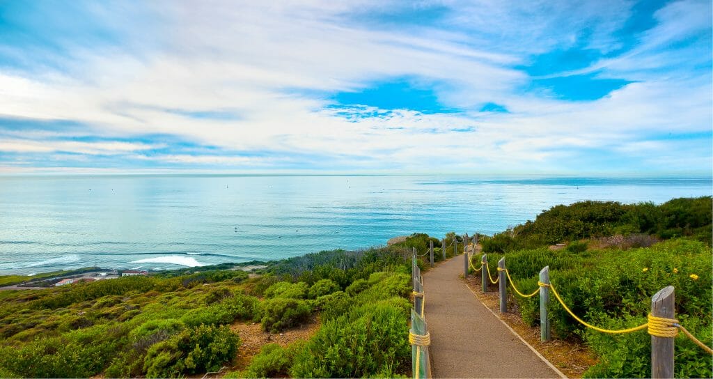 On a crystal clear day, the sky is bright blue and the ocean below it reflects its beauty while the hiking path curves to the right and the brush on either side becomes full and green