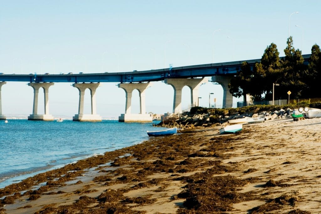 On the peaceful shores of the Coronado Bay, the water leisurely moves while cars zoom over the towering bridge in the distance in San Diego